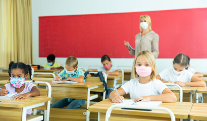 Fototapeta na wymiar Focused preteen schoolgirl in protective face mask studying in classroom. Necessary precautions in coronavirus pandemic