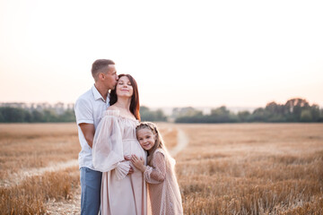 Young pregnant woman with her daughter and husband, happy family hugging on a golden field in the countryside. Family relations. Future parents