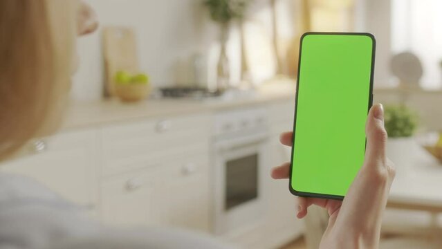 Back View of Young Woman at Home Sitting on Kitchen Room With Green Mock-up Screen Smartphone. Female is Watcing Content Without Touching Gadget Screen. Modent Technology