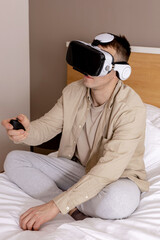 Obraz premium Young caucasian man sitting on bed at home with VR headset and playing interactive video game, exploring virtual reality. Man wearing VR glasses. Future, gadgets, technology, education, study online.