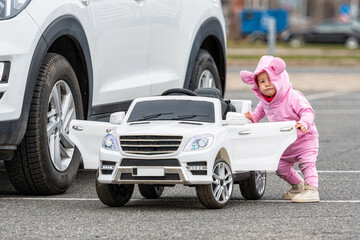a little girl at a big white childrens electric toy car in the parking lot next to a real car