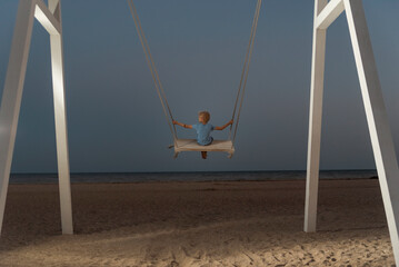 Little boy rides high in the sky on rope swing on sea background. Dark blue skies at twilight.