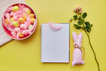 Easter mock up notepad. Pink bowl with colorful eggs, pink feathers, pink rose and handmade cute bunny. Happy Easter concept. Top view. Post card mock up on yellow background.