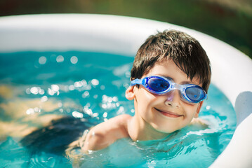 Little boy in swimming goggles is resting on edge of an inflatable circular pool outside on sunny summer day. Child is cheerful and happy. Close up. Summer vacation and healthy lifestyle concept.