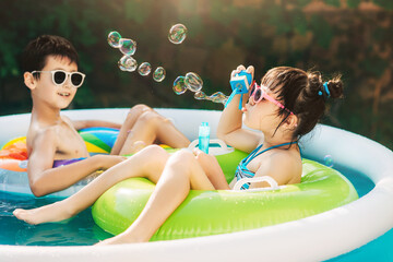 Little boy and girl, sibling, playing in inflatable pool outdoors swimming in inflatable circles on a sunny summer day. Girl blowing soap bubbles. Kids are fun and happy. Healthy lifestyle concept.