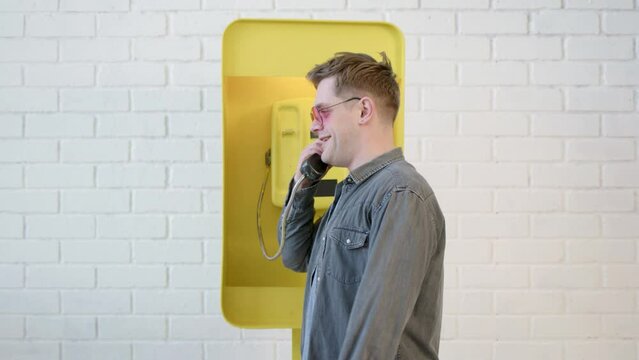Happy man with earring in his ear in pink glasses talking on phone in yellow telephone booth. Businessman on vacation talking to his business partners and smiling after hearing good news.