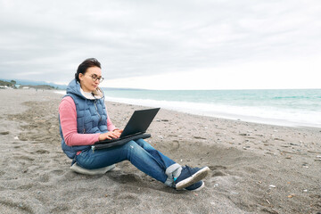 Beautiful sporty woman wearing eyeglasses with laptop computer working outdoors while sitting on winter beach in front of the sea. Modern lifestyle, connection, blogging, business, freelance work.