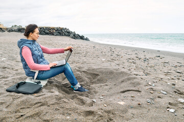 Beautiful sporty woman wearing eyeglasses with laptop computer working outdoors while sitting on winter beach in front of the sea. Modern lifestyle, connection, blogging, business, freelance work.