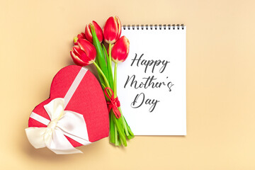 Bouquet of red tulips, gift box heart shaped, text Happy Mother's Day on open white notepad on beige background Top view Flat lay Holiday greeting card