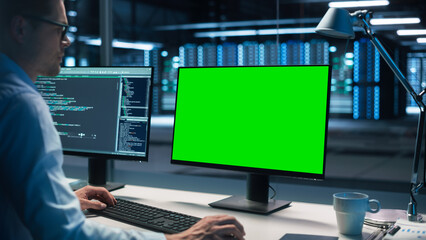 Male Specialist Working on Desktop Computer with Green Screen Mock Up Display in Evening Creative Office. Caucasian Information Specialist working at computer sitting at desk. Over the Shoulder Shot