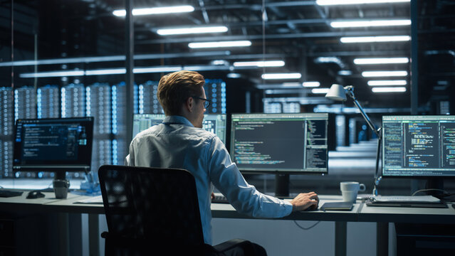 IT Specialist Uses Computer in Data Center. Server Farm Cloud Computing Facility with Male Maintenance Administrator Working During the Evening. Cyber Security and Network Protection.