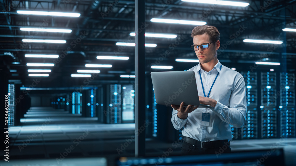 Poster caucasian young male it specialist using laptop, standing in data center. skilful man works on web s