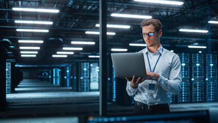 Caucasian Young Male IT Specialist Using Laptop, Standing in Data Center. Skilful Man Works on Web...