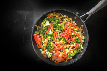 Vegetable pan with broccoli, red bell peppers and chickpeas on a black stove, healthy cooking for a vegan low carb diet to lose weight, copy space, high angle view from above