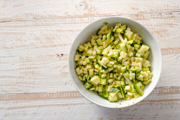 Zucchini cut into small cubes in a white bowl on a light rustic wooden table, healthy vegetable with minerals and vitamins but low calories, copy space, high angle view from above
