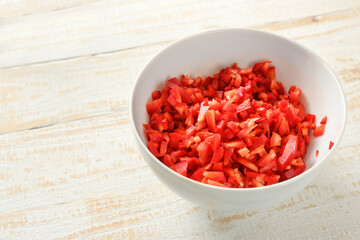 Red bell pepper cut into small cubes in a white bowl on a light rustic wooden table, healthy vegetable with vitamin c and capsaicin, copy space, selected focus