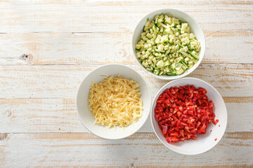 Diced vegetables, zucchini and red bell pepper, and grated cheese in white bowls on a rustic wooden...