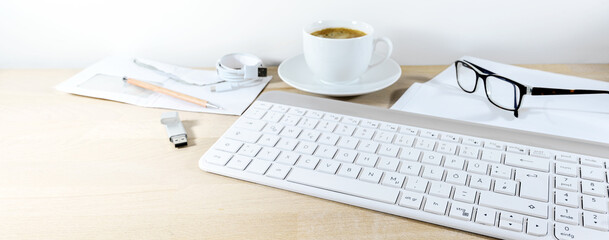 Light wooden office desk in panoramic format with computer keyboard, papers, glasses, usb stick and coffee cup, business at home, copy space, selected focus