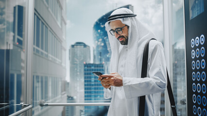 Successful Muslim Businessman in Traditional White Kandura Riding Glass Elevator to Office in Modern Business Center. Man Using Smartphone. Saudi, Emirati, Arab Businessman Concept.
