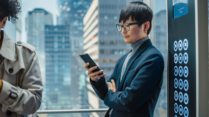 Successful Stylish Japanese Businessman Riding Glass Elevator to Office in Modern Business Center. Handsome Man Smile while Using Smartphone, Write Text Message, Check Social Media in a Lift. © Gorodenkoff