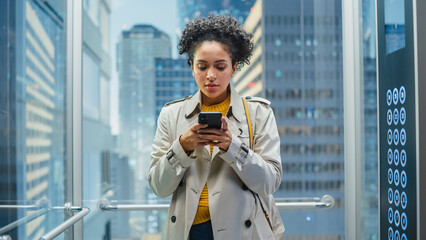 Beautiful Black Female Riding Glass Elevator to Office in Modern Business Center. Successful Manager Smile while Using Smartphone, Write Text Message, Check Social Media and Work Emails in a Lift. © Gorodenkoff