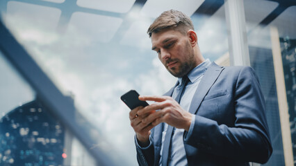 Successful Businessman Riding Glass Elevator to Office in Modern Business Center. Handsome Happy Man Focused while Using Smartphone, Write Text Message, Check Social Media and Work Emails in a Lift.