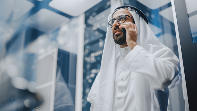 Successful Muslim Businessman in Traditional White Kandura Riding Glass Elevator to Office in Modern Business Center. Man Talking on a Phone in a Lift. Saudi, Emirati, Arab Businessman Concept.