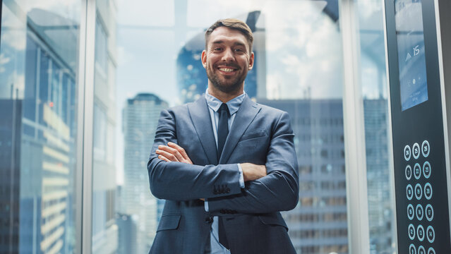 Portrait of a Successful Businessman in a Suit Riding Glass Elevator to Office in Modern Business Center. Male Looking at the Camera, Crossing Arms, Charmingly Smiling and Striking a Pose. - Powered by Adobe
