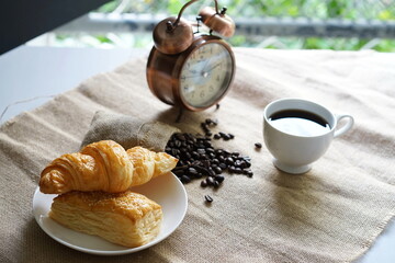 Croissants and pies are laid out on a white plate, with a white coffee mug, coffee beans packed in sacks, and an alarm clock on the side against a blurred natural background.
