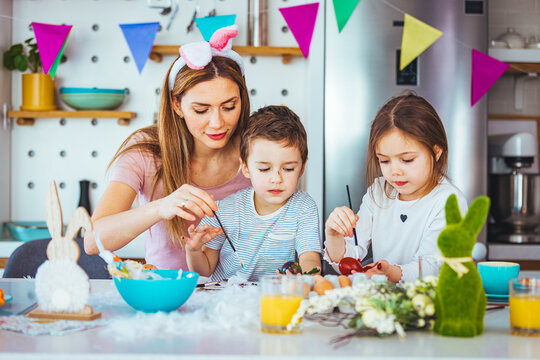 Joyful family wearing bunny ears headbands gathering at table in modern light kitchen and paining Easter eggs together. Happy family spending Easter together in the kitchen