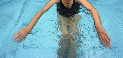 Schwimmerin mit schwarzem Badeanzug und ausgebreiteten Armen im Blauen Pool-Wasser am Abend im...