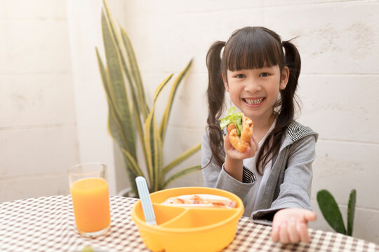 Happy Young Asian Girl Eating Breakfast And Orange Juice On The Table At Home. Healthy Eating, Food And Snacks, Ham And Cheese Sandwich