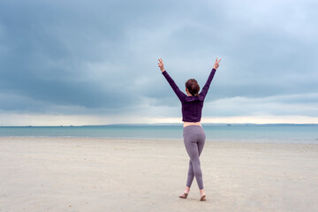 Obraz premium Young woman in sportswear is standing and lifting up arms for freedom on the beach, wearing fitnesswear