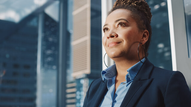 Portrait of Successful Black Businesswoman Riding Glass Elevator to Office in Modern Business Center. African American Female Looking at Modern Skyscrapers Out of the Panorama Window in the Lift.