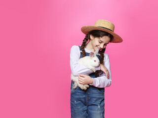 Little girl dressed in a jeans and straw hat holding a little white rabbit, isolated pink background.