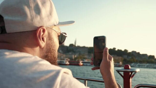 Close-up happy young bearded tourist man in eyeglasses takes smatphone picture of amazing summer sunset at cruise boat.