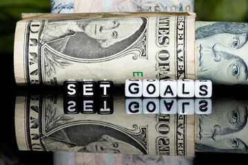 Mote alphabet blocks arranged into "set goals" on a background of rolls of banknotes.