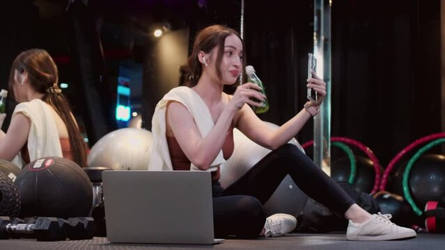 Young smiling woman at the gym relaxing and using mobile phone in fitness gym. Healthy lifestyle concept. Asian fit woman drinking vegetable water while video call with people at gym.