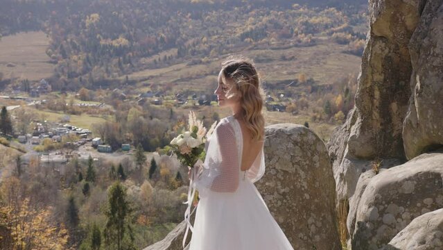 Pretty young bride wearing long white dress walking against mountains view. Natural sunset, rocks, hair in the wind.