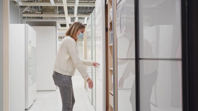a young woman in a protective mask in a furniture store chooses a tall wardrobe with pitched doors, taking out storage containers from it and inspecting them