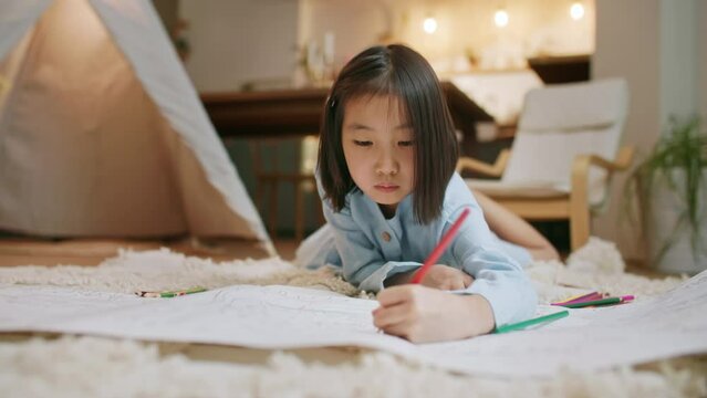 Talented little Asian Girl Paints on Paper, Using Color Pencil , Set Create a Colorful Emotional Painting With Paints. Preschool Girls are Engaged in Creativity at Home on Floor.