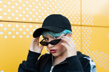 Portrait of a caucasian girl with a cap, short bluish hair and sunglasses looking at the camera