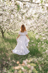Obraz na płótnie Canvas Fantasy woman in long white elegant fashion long dress walks in green spring blossom cherry garden. Happy cheerful girl princess bride. Skirt fabric flies flowing waving in wind motion.
