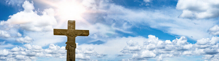 Religious crucifixion Easter background banner panorama - Old stone cross crucifix with blue sky, clouds and sunbeams..