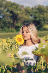 Beautiful young long-haired blonde woman in a white blouse with bare shoulders holds a sunflower in her hands on a sunflower field.Summer concept.