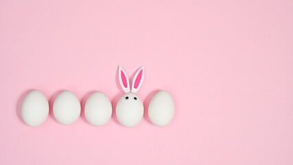 Creative composition with white eggs and one with rabbit ears on pastel pink background. FLat lay creative Easter copy space.