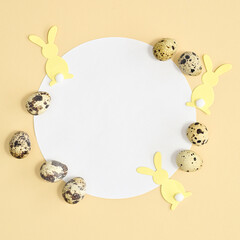 Round paper card copy space with yellow rabbits and natural quail eggs on pastel beige background. Flat lay Easter concept