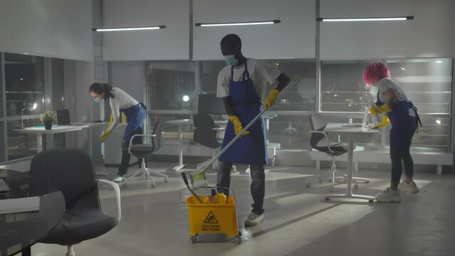 Diverse team of janitors in facemask clean empty office and leave