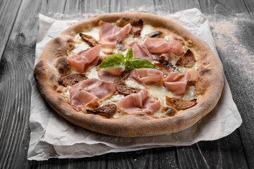 Delicious freshly baked meat pizza. Appetizing whole-wheat pizza with cheese, prosciutto and figs. Served on parchment paper. Dark wooden background with sprinkled flour.