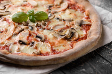 Freshly baked meat pizza. Appetizing pizza from whole grain dough with cheese, mushrooms and chicken meat. Served on parchment paper. Dark wooden background with sprinkled flour. Cropped image.